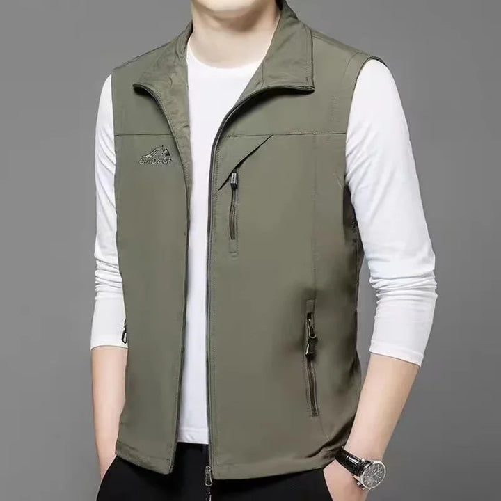 Mann trägt olivgrüne Weste mit Reißverschlusstaschen über weißem Langarmshirt, grauer Hintergrund. Outdoor-Mode, Herrenbekleidung, Freizeitlook.