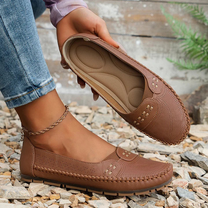 Braune Damen-Mokassins aus Leder mit Ziernähten, perforiertem Design und bequemer Sohle, getragen mit Jeans auf Kieselsteinen.