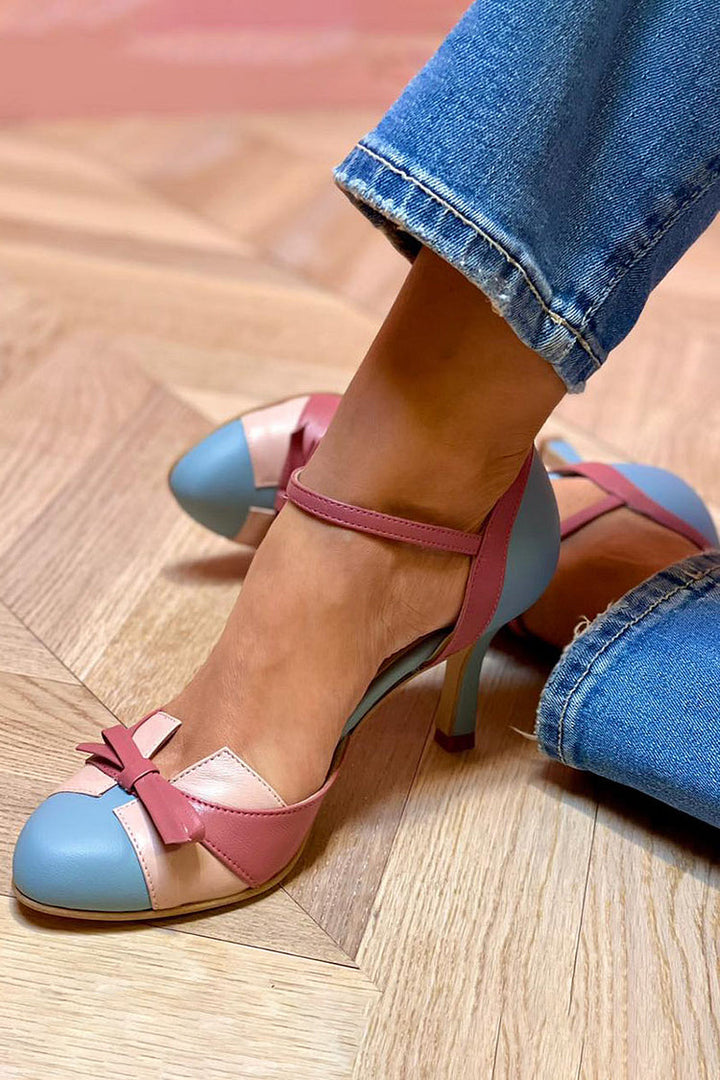 Frau trägt elegante, zweifarbige High Heels in Rosa und Blau mit Schleifen-Detail, kombiniert mit blauen Jeans. Modische Damenschuhe auf Holzboden.