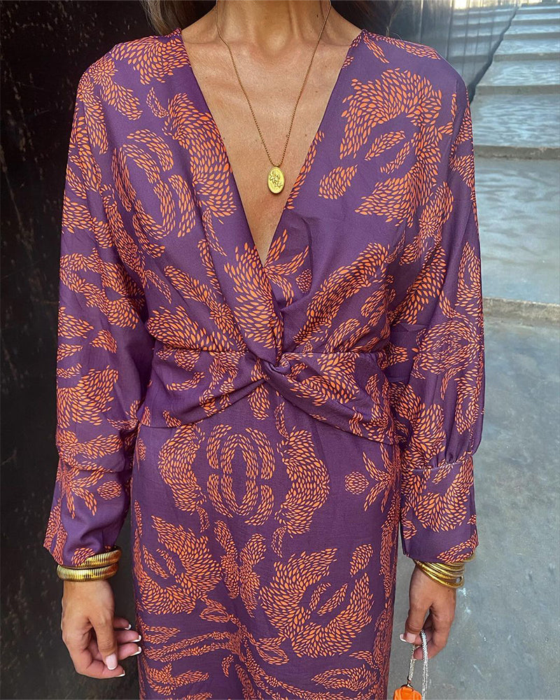 Frau in lila Kleid mit orangefarbenem Muster, V-Ausschnitt und goldenen Accessoires. Elegantes Outfit für Abendveranstaltungen.
