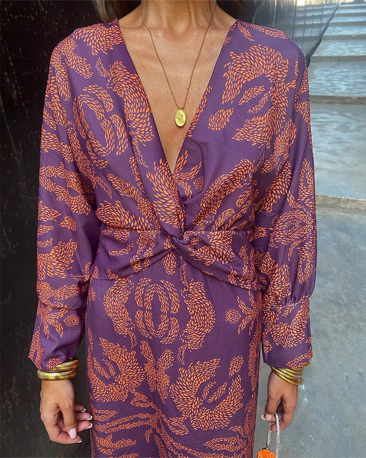 Frau in lila Kleid mit orangefarbenem Muster, V-Ausschnitt und goldenen Accessoires. Elegantes Outfit für Abendveranstaltungen.
