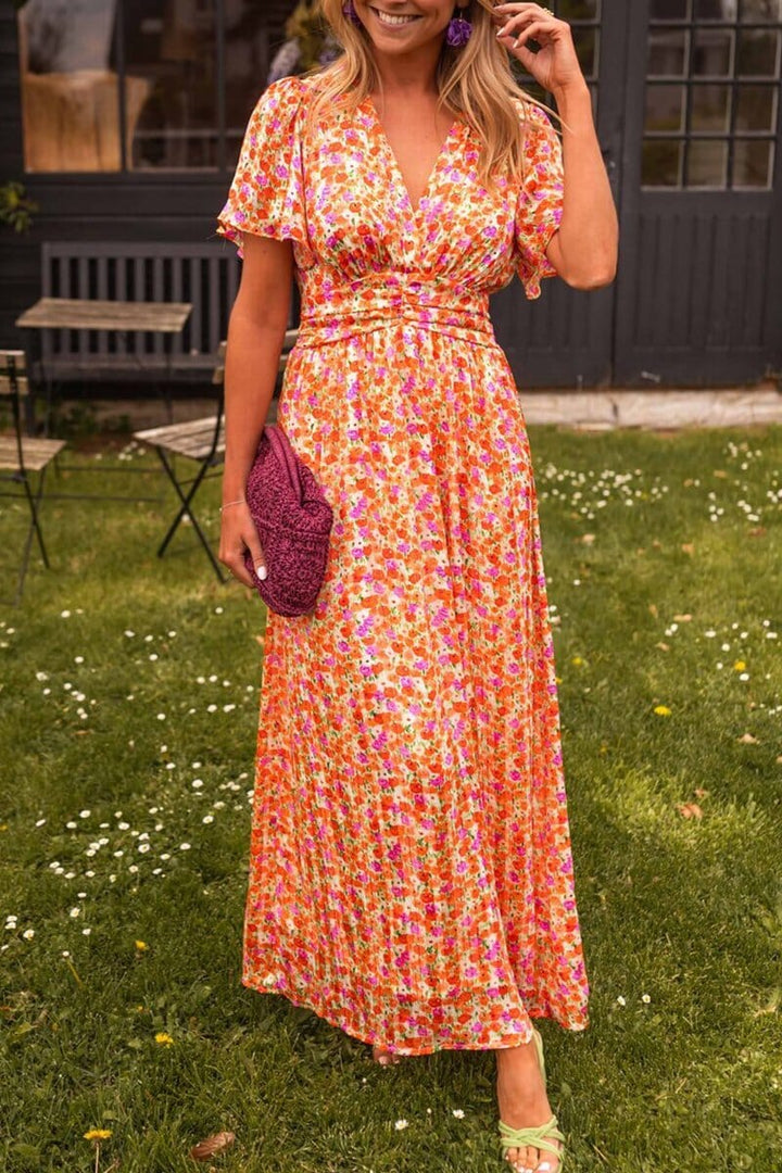 Frau in langem, geblümtem Sommerkleid mit lila Tasche steht auf Rasen vor dunklem Gebäude. Modetrend, Sommermode, Damenkleid, Blumenmuster.