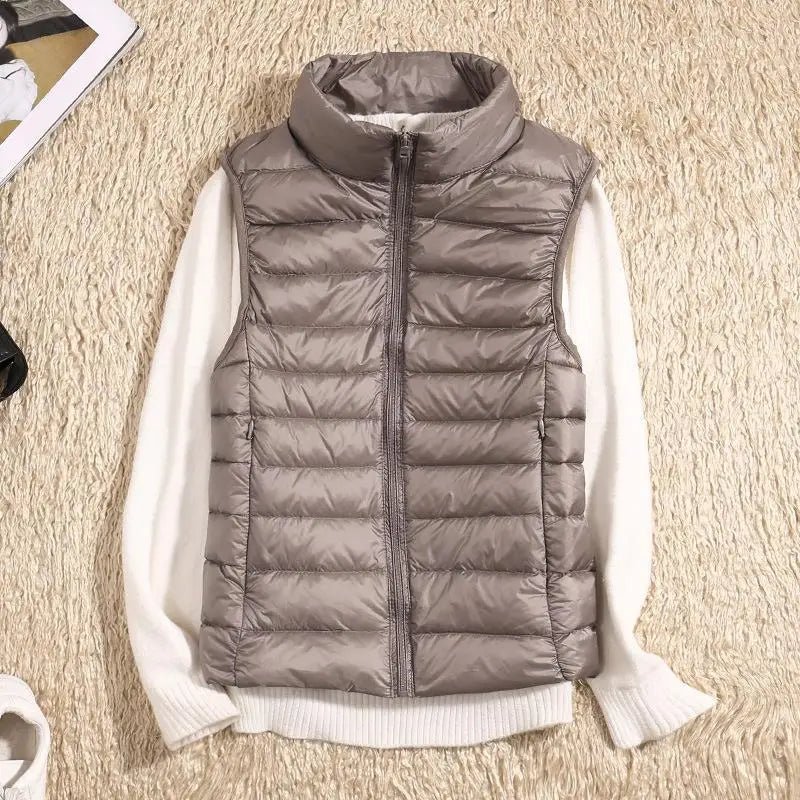 Graue Steppweste mit Reißverschluss auf cremefarbenem Pullover, flach auf beigem Teppich liegend. Modische Herbstbekleidung, Damenmode.