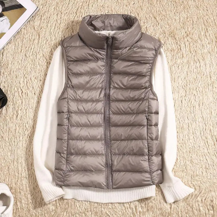 Graue Steppweste mit Reißverschluss auf cremefarbenem Pullover, flach auf beigem Teppich liegend. Modische Herbstbekleidung, Damenmode.