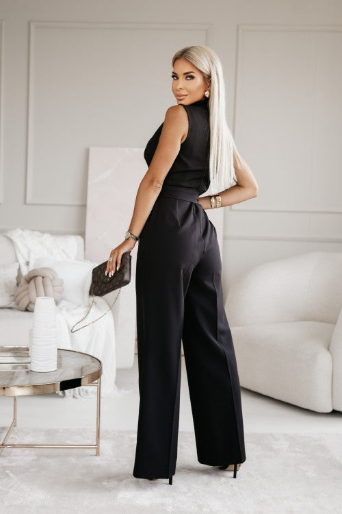 Frau in eleganter schwarzer Abendmode mit langer blonder Frisur, hält eine kleine Handtasche, steht in modernem Wohnzimmer mit stilvoller Einrichtung.