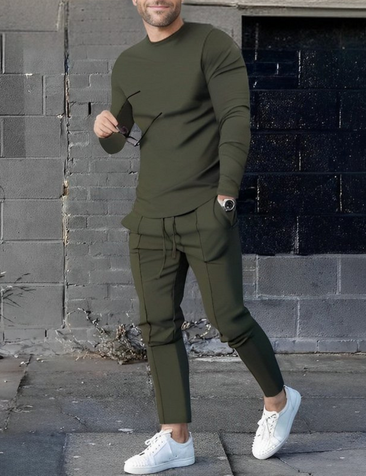 Mann in olivgrünem Freizeitanzug mit Sonnenbrille und weißen Sneakers vor grauer Backsteinmauer. Modetrend, Herrenmode, urbaner Stil.