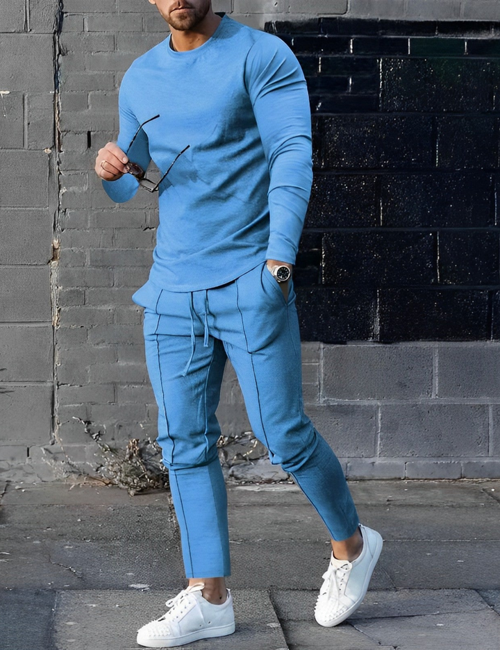 Mann in blauem Trainingsanzug mit Sonnenbrille und weißen Sneakers vor grauer Wand. Modischer Freizeitlook, Streetwear, Herrenmode.