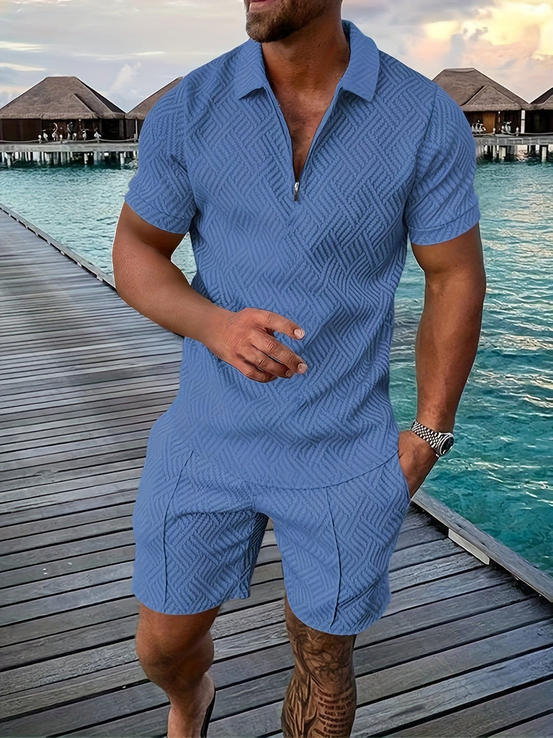 Mann in blauem Sommeroutfit auf Holzsteg vor Wasserbungalows, tropische Kulisse, stilvolle Freizeitkleidung, Urlaubsstimmung, Mode am Meer.