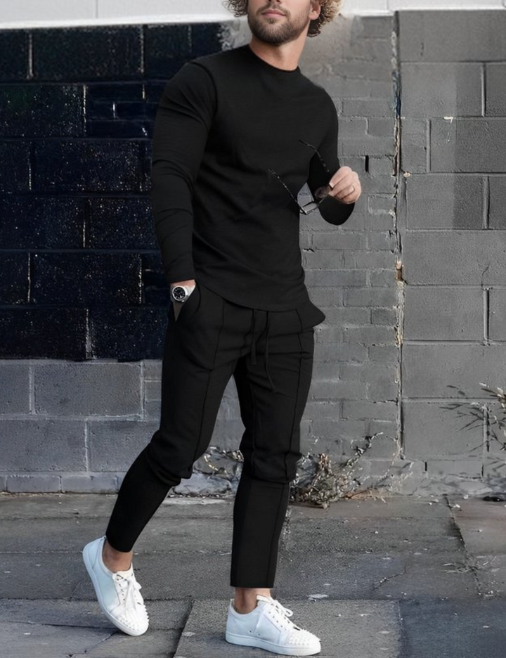 Mann in schwarzem Outfit mit weißem Sneaker, hält Sonnenbrille, steht vor grauer Ziegelwand. Streetwear, Mode, Herrenbekleidung, urbaner Stil.