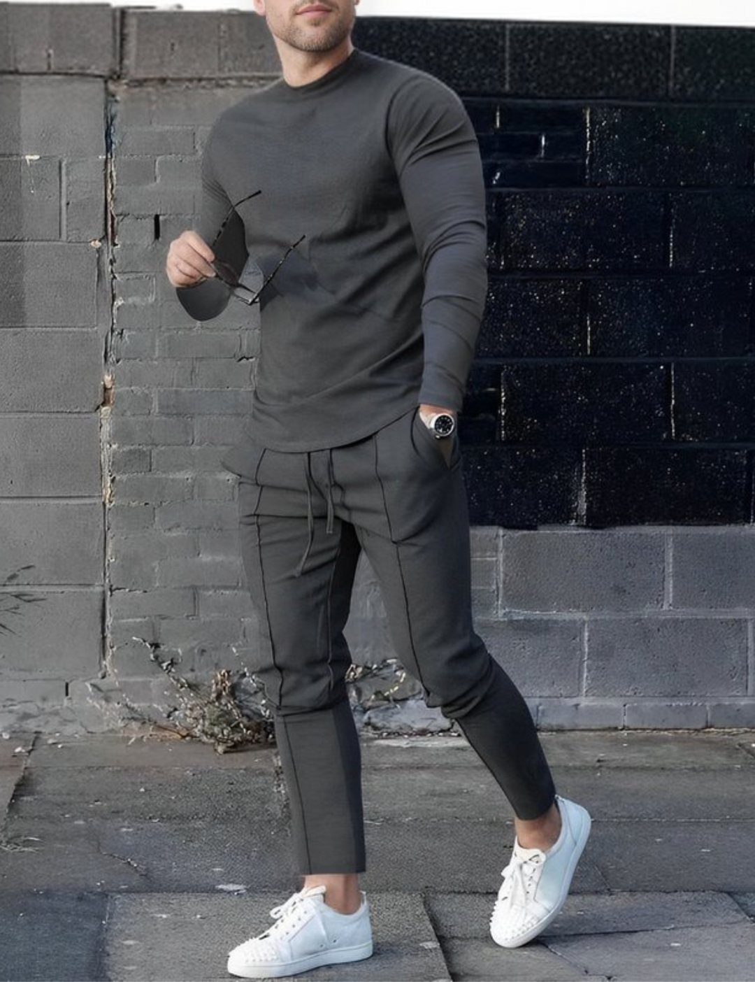 Mann in grauem Freizeitanzug mit Sonnenbrille und weißen Sneakers vor grauer Backsteinmauer. Modetrend, Herrenmode, urbaner Stil.
