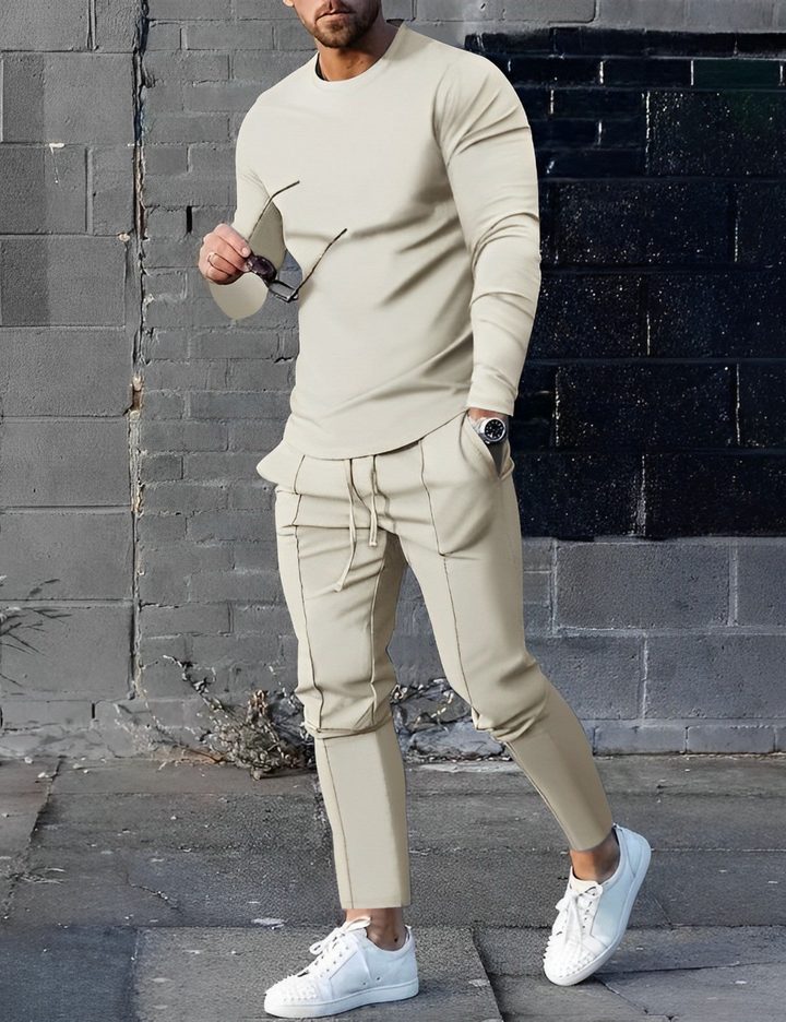 Mann in beigem Jogginganzug mit Sonnenbrille und weißen Sneakers vor grauer Backsteinmauer. Streetwear, Herrenmode, lässiger Stil.