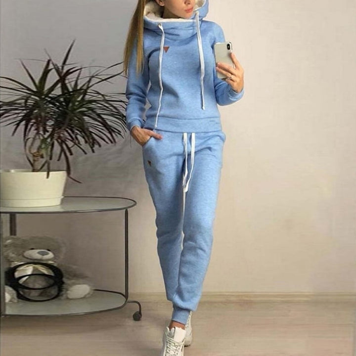 Frau in hellblauem Jogginganzug mit Kapuze, steht neben einem Tisch mit Pflanze und Plüschtier, Smartphone in der Hand. Bequeme Freizeitmode.
