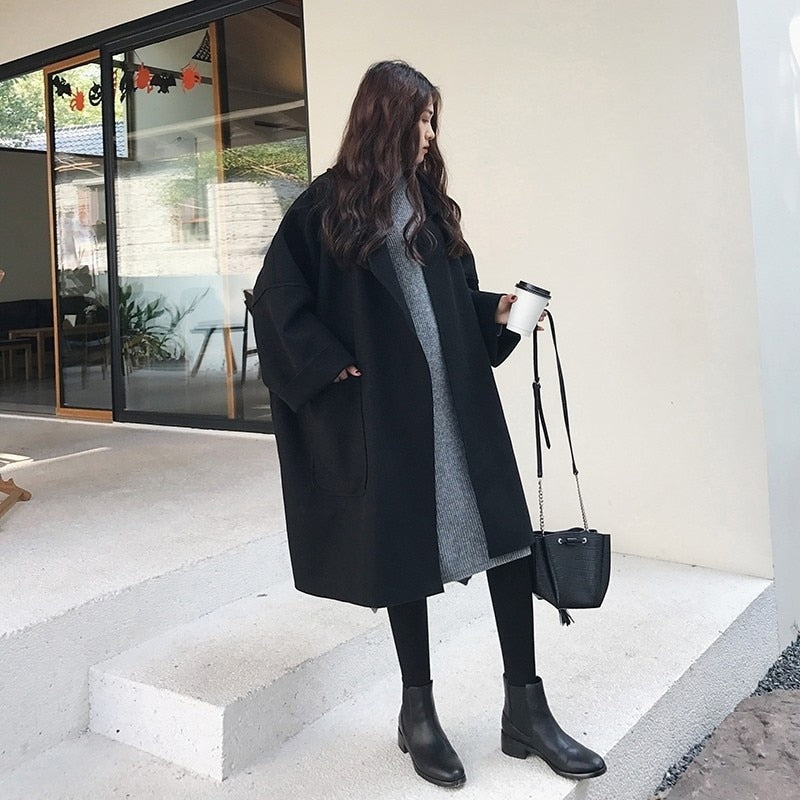 Frau in schwarzem Mantel und grauem Kleid, hält Kaffeebecher, steht vor modernem Gebäude. Herbstmode, Streetstyle, lässiger Look.