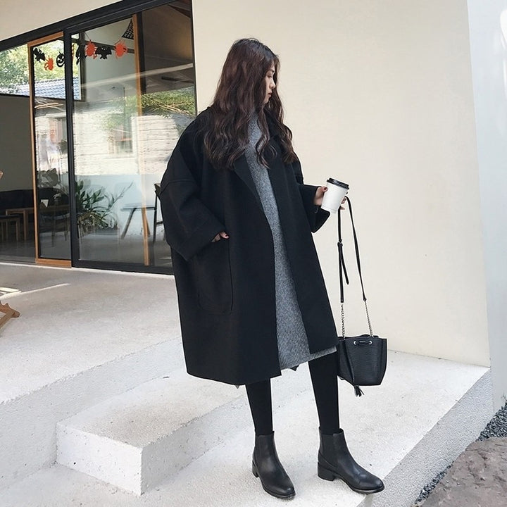 Frau in schwarzem Mantel und grauem Kleid, hält Kaffeebecher, steht vor modernem Gebäude. Herbstmode, Streetstyle, lässiger Look.
