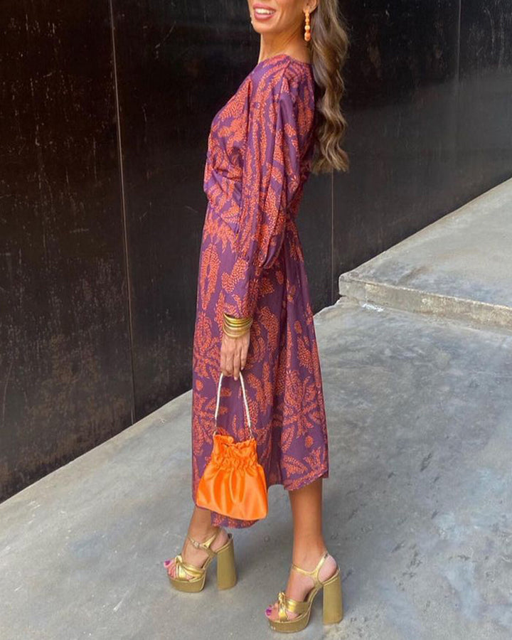 Frau in lila-orange gemustertem Kleid, trägt orange Tasche und goldene Plateau-Sandalen. Elegante Mode, Streetstyle, Sommeroutfit.