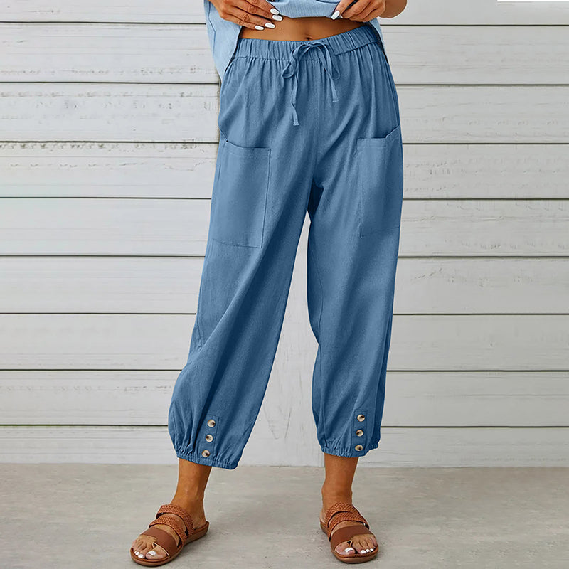 Frau trägt blaue Leinenhose mit Taschen und Knöpfen am Saum, kombiniert mit Sandalen. Sommermode, lässiger Stil, bequeme Damenbekleidung.