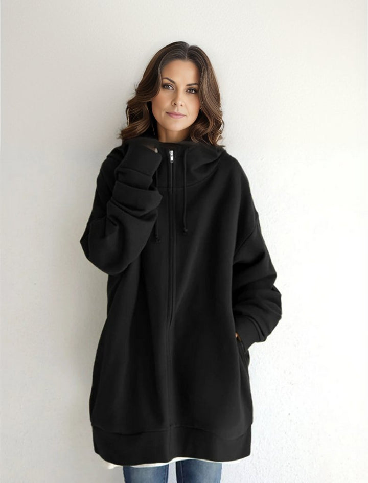 Eine Frau in einem übergroßen schwarzen Hoodie mit Reißverschluss und Kapuze, lässig vor einer weißen Wand stehend. Mode, Streetwear, Damenbekleidung.