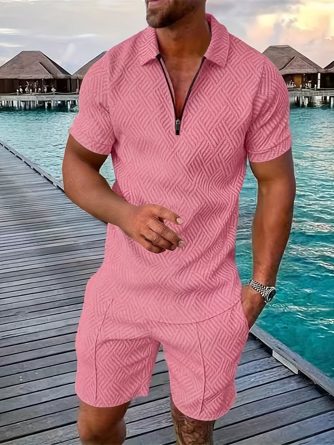 Mann in rosa Sommeranzug auf Holzsteg am Meer, tropische Bungalows im Hintergrund, stilvolle Freizeitmode, Urlaubskleidung, Herrenmode.