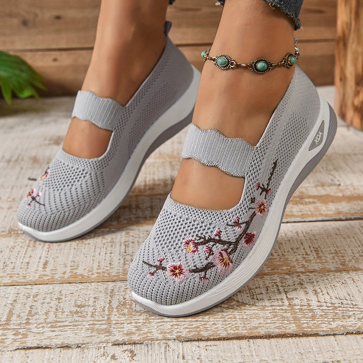 Graue Damen-Sneaker mit Blumenstickerei, atmungsaktivem Mesh-Obermaterial und Keilabsatz, getragen auf Holzboden. Perfekt für Frühling und Sommer.