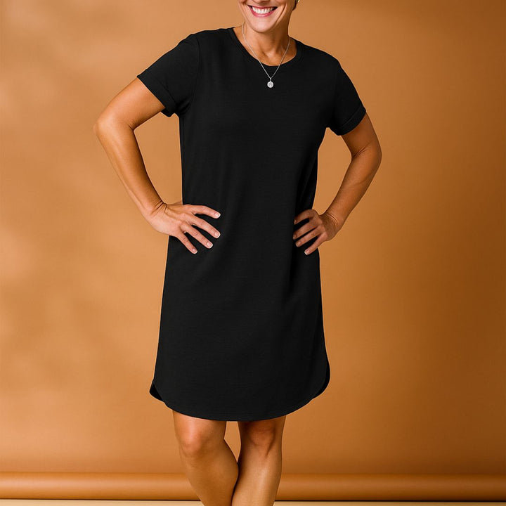 Frau in schwarzem T-Shirt-Kleid vor braunem Hintergrund, lächelnd, mit Händen in der Hüfte. Modetrend, lässiges Damenoutfit, Sommerkleidung.