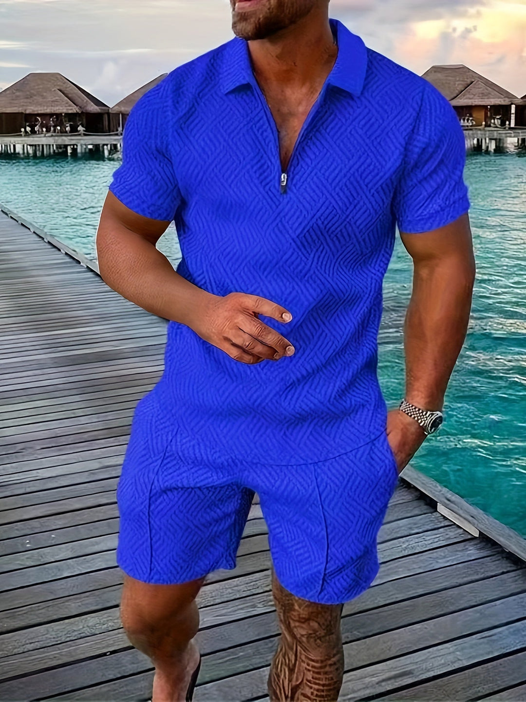 Mann in blauem Sommeroutfit auf Holzsteg am Meer, im Hintergrund Wasserbungalows. Mode, Herrenbekleidung, Urlaub, tropische Kulisse.