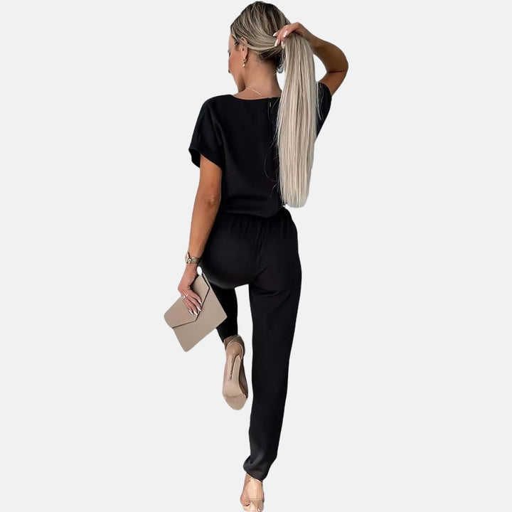 Frau in schwarzem Jumpsuit, hält beige Clutch, zeigt Rückenansicht. Elegante Mode, lange blonde Haare, hohe Absätze. Ideal für Abendveranstaltungen.