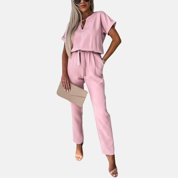 Frau in rosa Jumpsuit mit Gürtel, hält beigefarbene Clutch, trägt Sonnenbrille und Sandalen. Modetrend, Damenmode, eleganter Look.