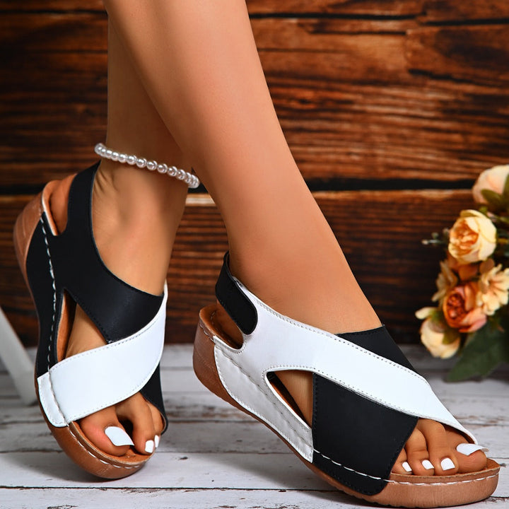 Damen Sandalen in Schwarz-Weiß mit Keilabsatz, Kreuzriemen-Design und Perlenfußkettchen, auf Holzhintergrund mit Blumen. Ideal für Sommermode.