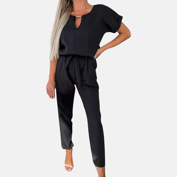 Frau in schwarzem Jumpsuit mit kurzen Ärmeln und V-Ausschnitt, modisch elegant, stehend vor weißem Hintergrund, Sommermode Damenbekleidung.