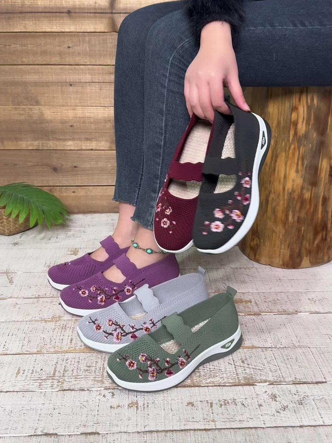 Frau hält drei Paar bestickte Damen-Sneaker in Lila, Rot und Schwarz. Schuhe auf Holzboden, modisch und bequem. Ideal für Freizeitkleidung.