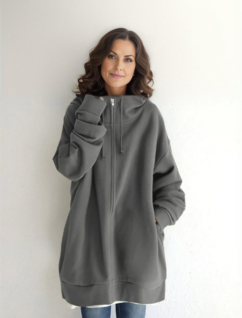 Eine Frau in einem übergroßen grauen Hoodie mit Reißverschluss und Kapuze, lächelt vor einem weißen Hintergrund. Bequeme Freizeitmode, lässiger Stil.