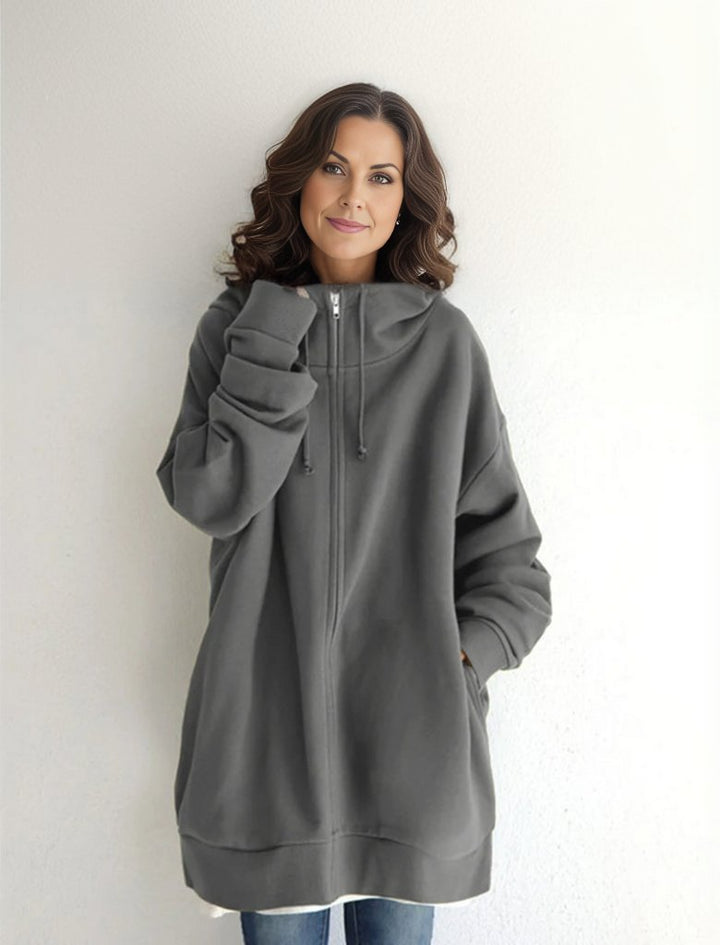 Eine Frau in einem übergroßen grauen Hoodie mit Reißverschluss und Kapuze, lächelt vor einem weißen Hintergrund. Bequeme Freizeitmode, lässiger Stil.