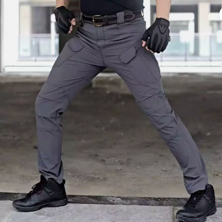 Mann in taktischer Hose, graue Outdoor-Hose mit Taschen, schwarze Handschuhe und Stiefel, robuste Kleidung für Abenteuer und Outdoor-Aktivitäten.