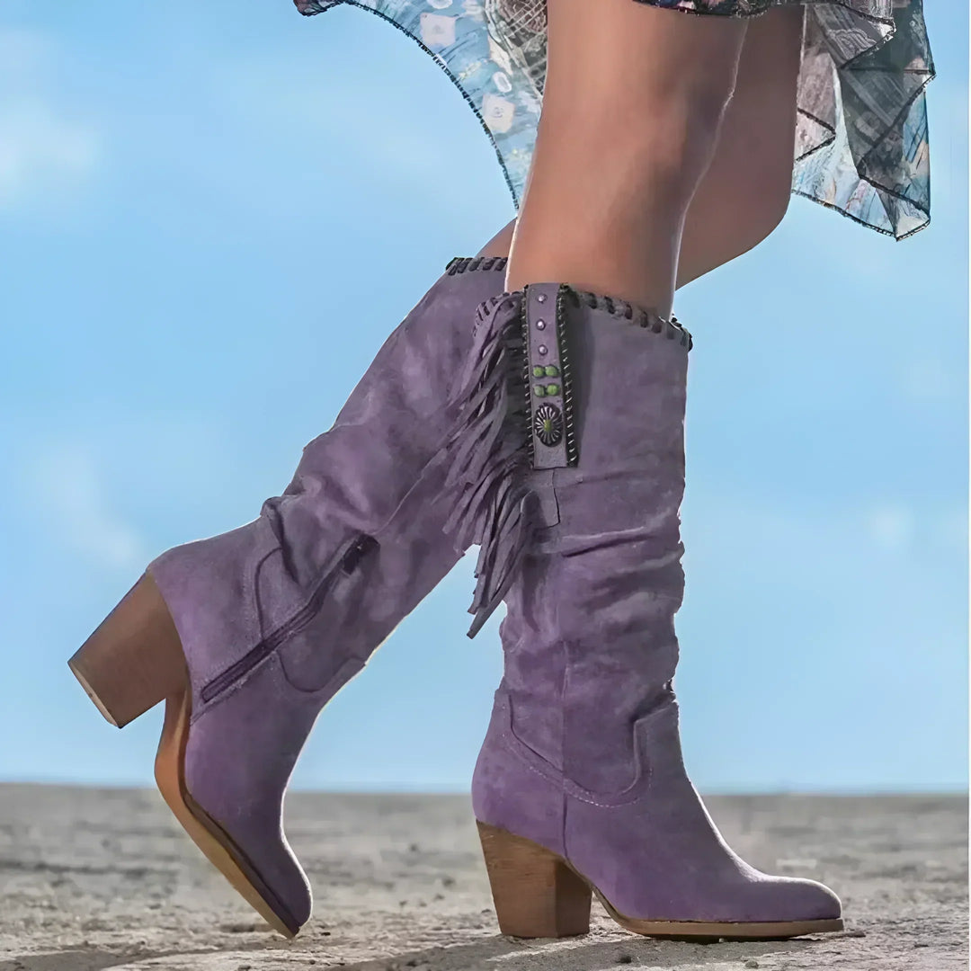 Lila Cowboystiefel mit Fransen und Absatz auf sandigem Boden, Damenmode, Westernstil, trendige Stiefel, Outdoor-Fashion, blauer Himmel im Hintergrund.