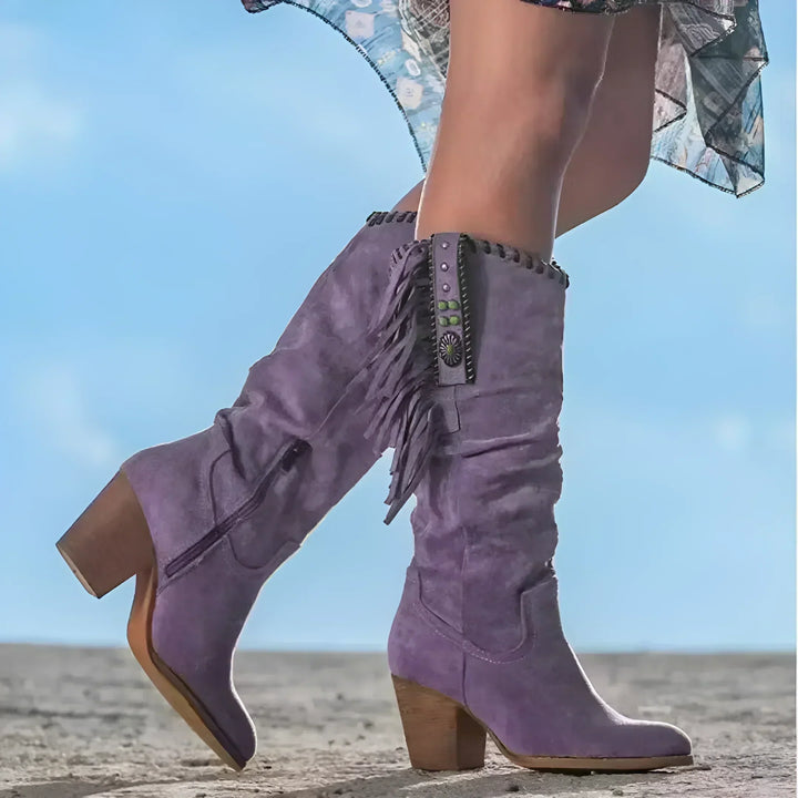 Lila Cowboystiefel mit Fransen und Absatz auf sandigem Boden, Damenmode, Westernstil, trendige Stiefel, Outdoor-Fashion, blauer Himmel im Hintergrund.