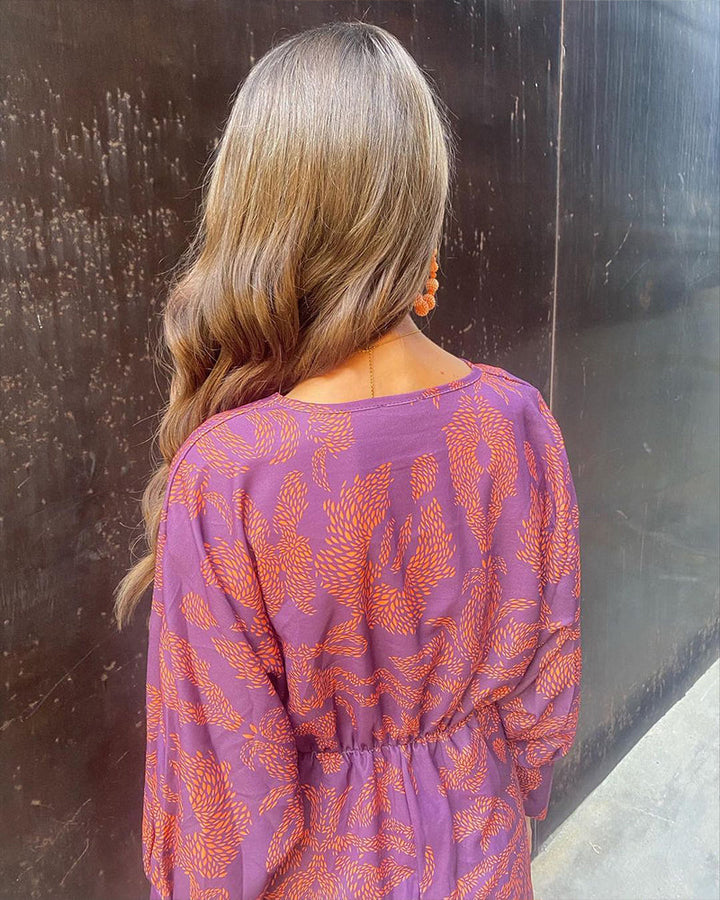 Frau mit langen, welligen Haaren in lila Kleid mit orangefarbenem Muster, steht vor dunkler Wand. Modetrend, Damenmode, Herbstoutfit.