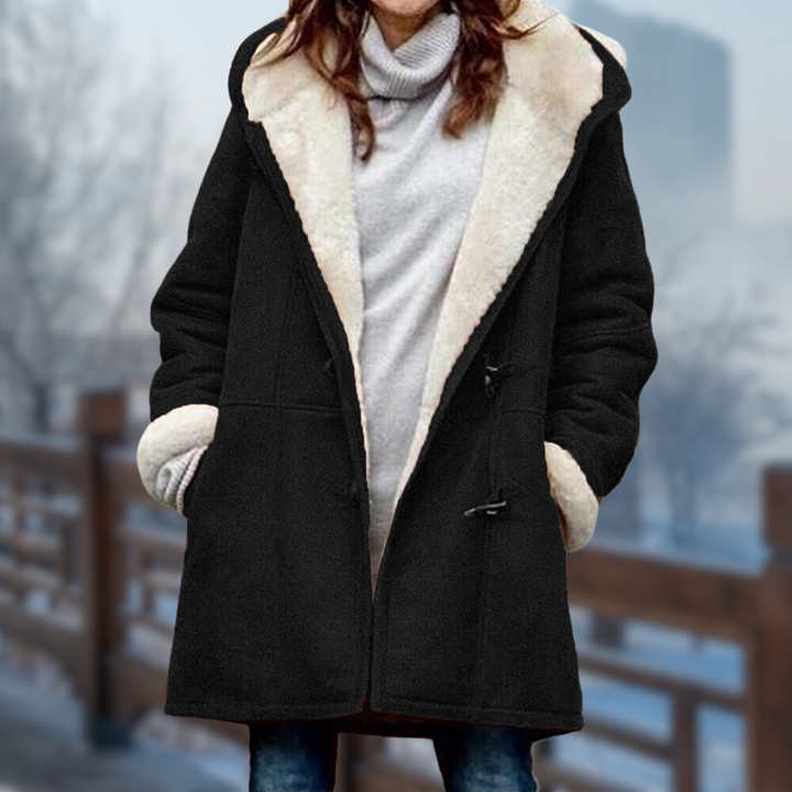 Frau in schwarzem Wintermantel mit weißem Fellkragen, grauem Rollkragenpullover und blauer Jeans steht auf einer Brücke im Winter.