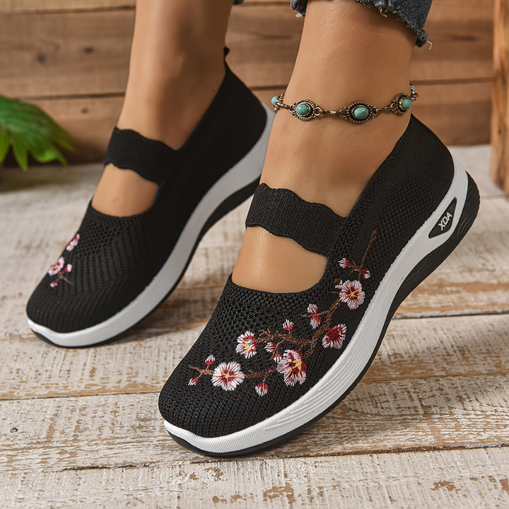 Schwarze Damen-Sneaker mit Blumenstickerei, bequeme Slip-On-Schuhe, weiße Sohle, ideal für Freizeit und Mode, getragen mit Jeans und Fußkettchen.
