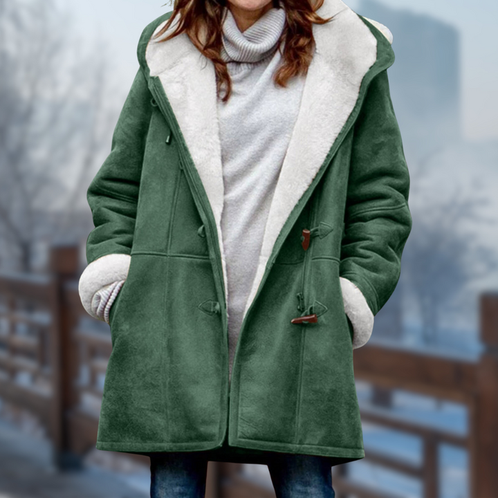 Frau in grünem Wintermantel mit Kapuze und Knebelknöpfen, steht auf einer verschneiten Brücke. Wintermode, Damenmantel, Outdoor-Bekleidung.