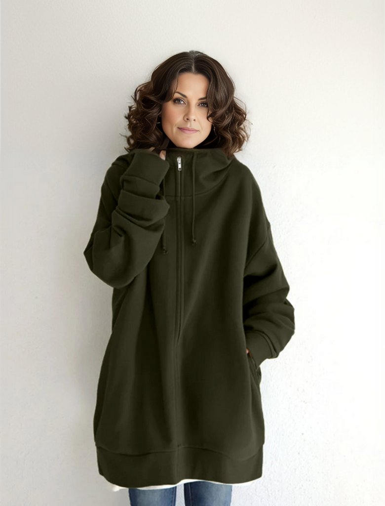 Frau in übergroßem, olivgrünem Hoodie mit Reißverschluss und Kapuze, lässiger Stil, weiße Wand im Hintergrund, modische Freizeitkleidung.