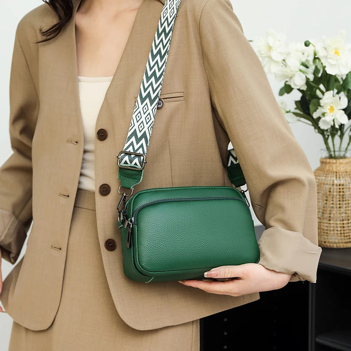 Frau in beigem Anzug mit grüner Ledertasche und gemustertem Schulterriemen, vor einem Blumenstrauß. Elegante Damenmode, Accessoires.