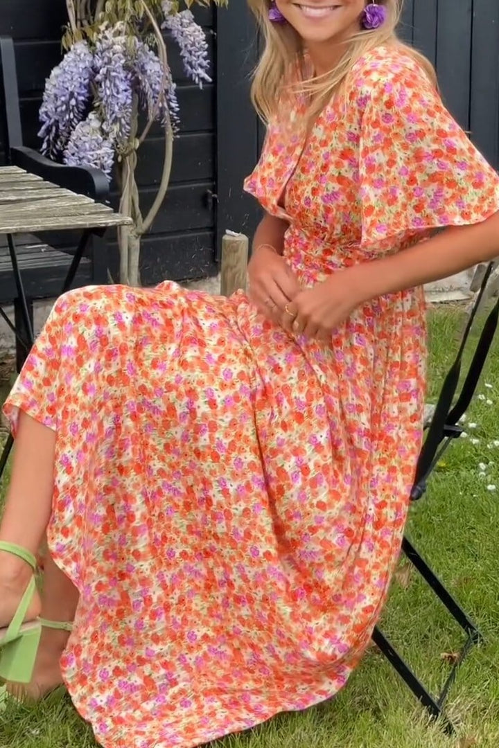 Frau in geblümtem Sommerkleid sitzt auf Stuhl im Garten, trägt grüne Sandalen und lila Ohrringe, umgeben von blühenden Pflanzen.