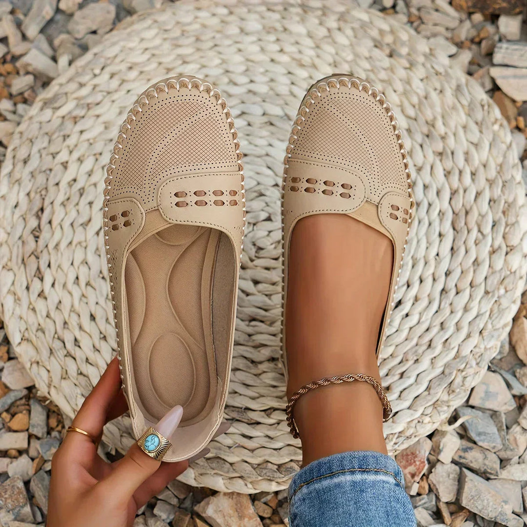 Beige Damen-Loafer aus Leder mit Lochmuster, getragen auf einem runden, geflochtenen Teppich, umgeben von Kieselsteinen. Perfekt für Sommermode.
