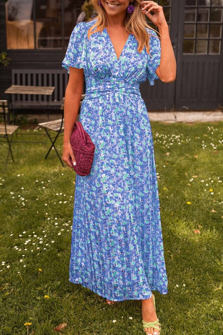 Frau in blauem, geblümtem Maxikleid mit violetter Clutch auf Rasen vor dunklem Gebäude. Sommermode, elegantes Outfit, Freizeitkleidung.
