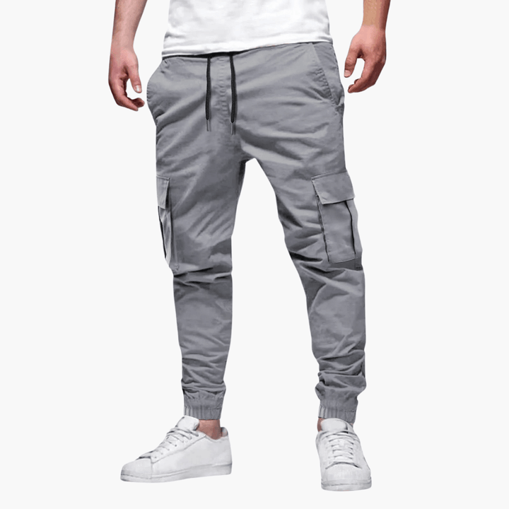 Graue Cargo-Jogginghose für Herren, lässiger Stil, mit Seitentaschen und Kordelzug, kombiniert mit weißen Sneakers, ideal für Freizeitmode.