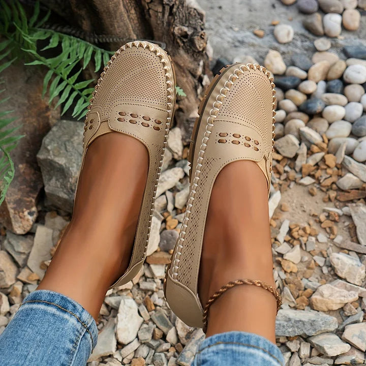 Beige Damen-Loafer aus Leder mit Lochmuster, getragen von einer Person in Jeans, auf einem steinigen Untergrund. Bequeme Sommerschuhe für Frauen.
