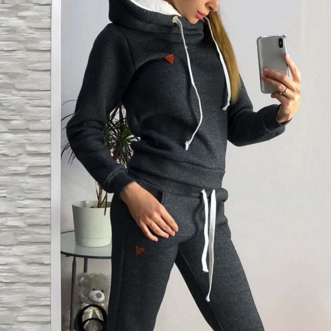Frau im grauen Jogginganzug mit Kapuze macht Selfie, moderne Freizeitmode, gemütlicher Hoodie, Smartphone, Indoor-Pflanze im Hintergrund.