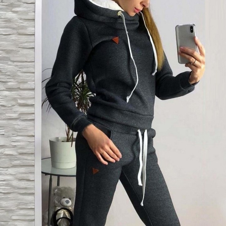 Frau im grauen Jogginganzug mit Kapuze macht Selfie, moderne Freizeitmode, gemütlicher Hoodie, Smartphone, Indoor-Pflanze im Hintergrund.