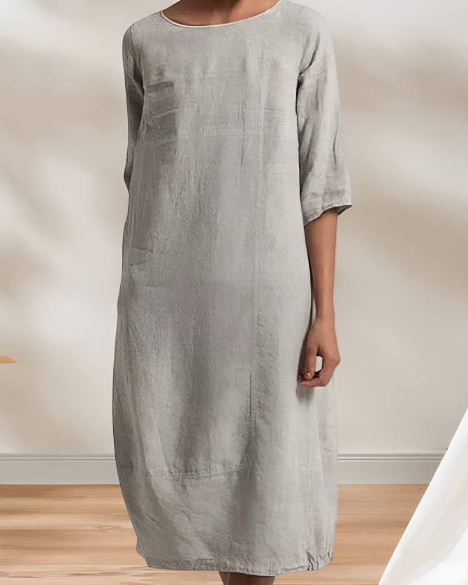 Frau in grauem Leinenkleid, knielang, mit 3/4-Ärmeln, steht auf Holzboden vor neutralem Hintergrund. Minimalistisches, elegantes Design.