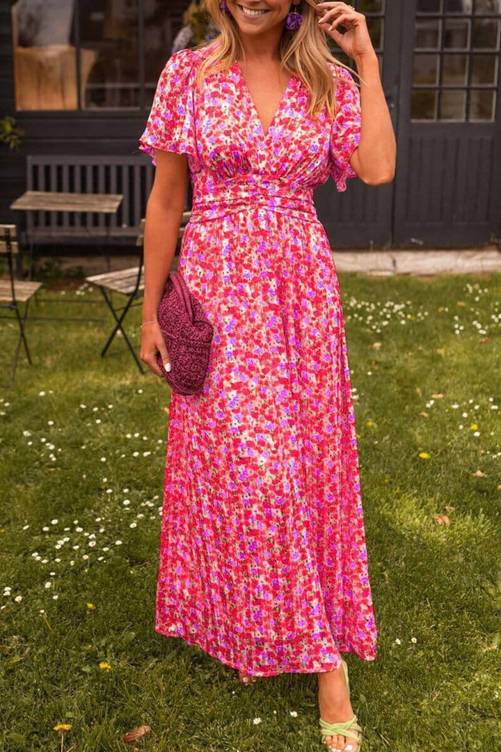 Frau in langem, pinkem Blumenkleid mit Clutch auf Rasen vor dunklem Gebäude. Sommermode, florales Design, elegantes Outfit.